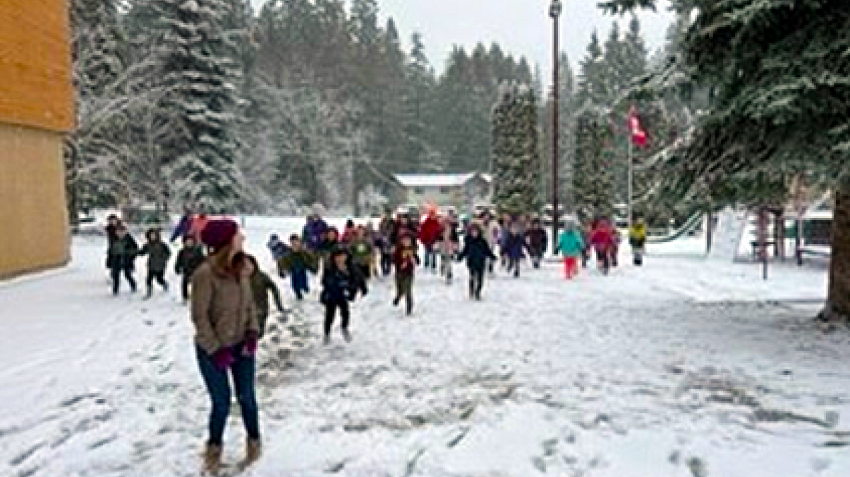 A young, female adult wearing a burgundy toque leading school children in play in a snowy winter schoolyard