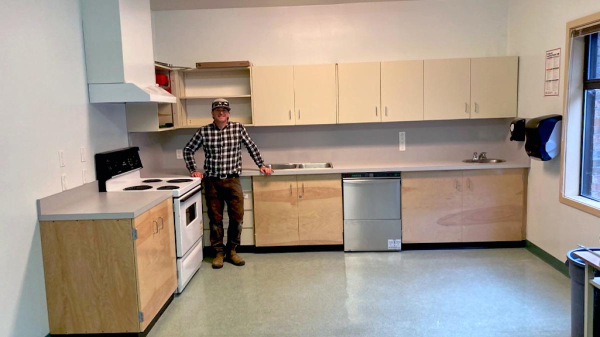 A man in brown pants, a black plaid shirt and a ballcap with sunglasses on the brim stands in front of a kitchen counter in a brand new kitchen