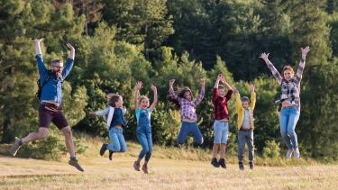 People in a field in front of a forest jumping in the air with their hands up