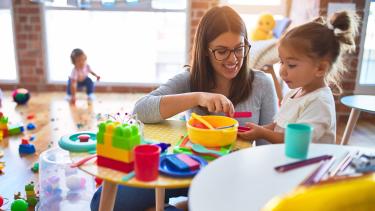 Woman with brown hair and glasses helps a little girl play at a table.